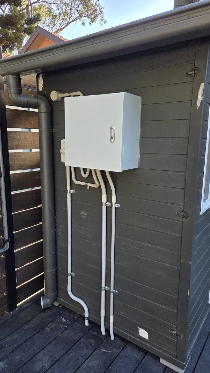 White electrical enclosure mounted on an exterior wall with rigid and flexible conduit running down to a deck